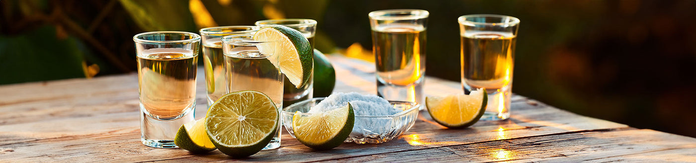 Sechs Tequila-Shots mit Limettenspalten und Salz auf einem Holztisch im warmen Sonnenlicht.