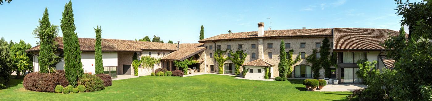 Eine große, rustikale Villa mit Steinmauern, Bogenfenstern und Ziegeldächern, umgeben von Bäumen und grünen Rasenflächen.