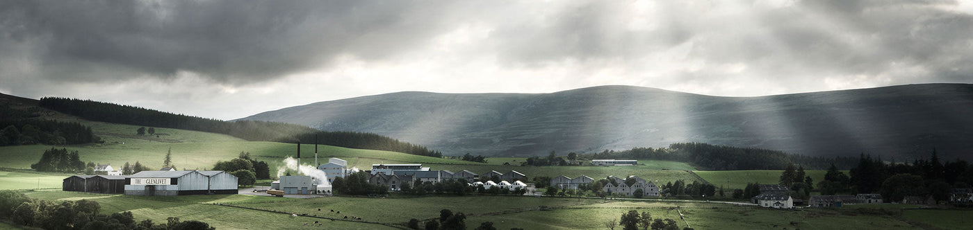 Glenlivet Whisky Destillerie in Schottland vor grünen Hügeln und dramatischem Himmel mit Sonnenstrahlen