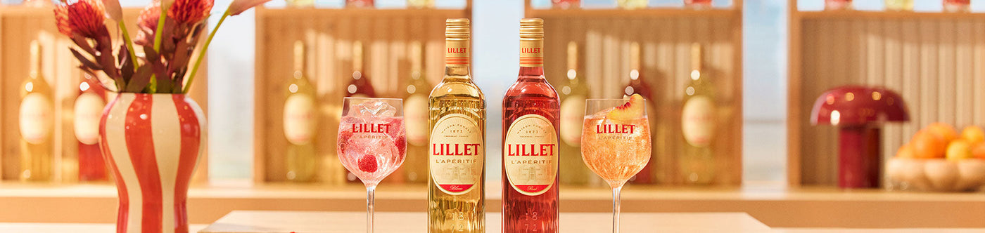 Zwei Flaschen Lillet mit passenden Aperitif-Gläsern, serviert auf einer stilvollen Bar mit Deko und Früchten