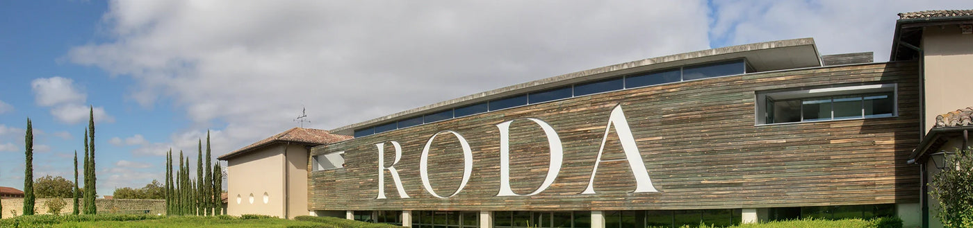 Modernes Gebäude mit großen RODA-Buchstaben an der Holzfassade, umgeben von Grünflächen und hohen Bäumen unter einem bewölkten Himmel.