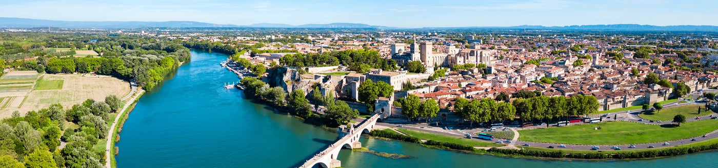 Luftaufnahme einer historischen Stadt mit einem Fluss, einer Brücke, alten Gebäuden und grünen Bäumen unter einem strahlend blauen Himmel.