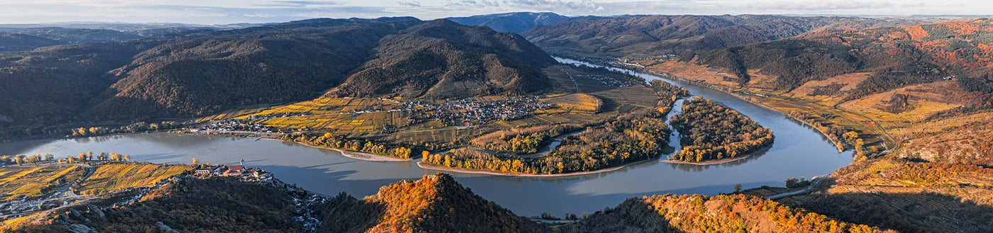 Ein breiter Fluss schlängelt sich durch ein Tal mit Hügeln, Wäldern und einem Dorf, das von herbstlichem Laub umgeben ist.